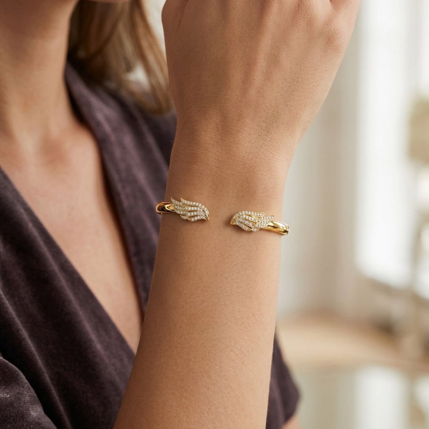 A close-up of a woman's wrist wearing a gold cuff bracelet with pavé-set clear stones and a small gold accent on each side, shown against a softly blurred indoor background.