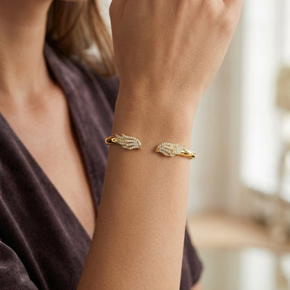 A close-up of a woman's wrist wearing a gold cuff bracelet with pavé-set clear stones and a small gold accent on each side, shown against a softly blurred indoor background.