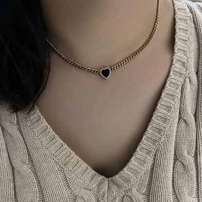 A close-up of a person wearing a gold chain necklace with a small black heart-shaped pendant, paired with a beige cable-knit sweater.