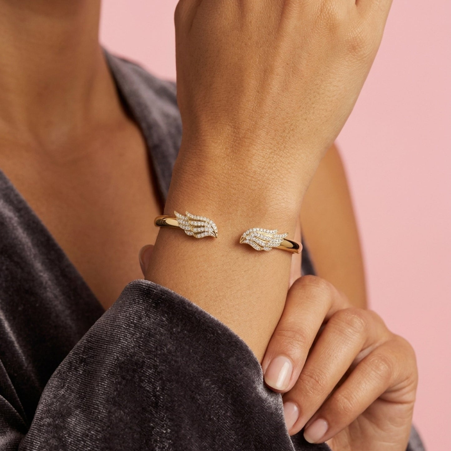 A close-up of a person's wrist wearing a gold cuff bracelet with two wing-shaped settings adorned with small clear stones, set against a soft pink background.