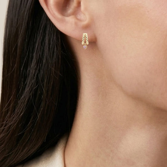 Close-up of a model wearing a small gold hoop earring with a heart gemstone, paired with a silver chain necklace and a cream-colored top.