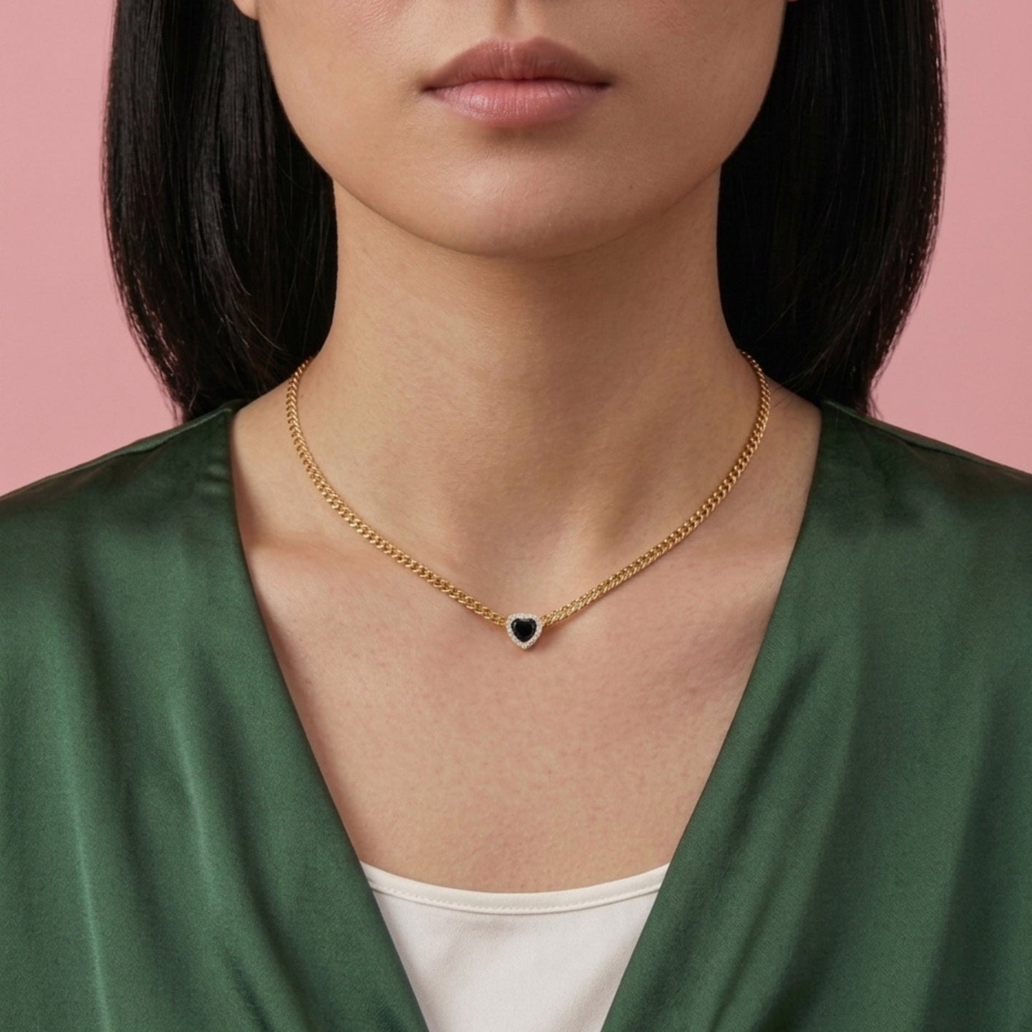 A close-up of a model wearing a delicate gold chain necklace with a small heart-shaped pendant featuring a dark blue center, styled with a green satin top against a solid pink background.