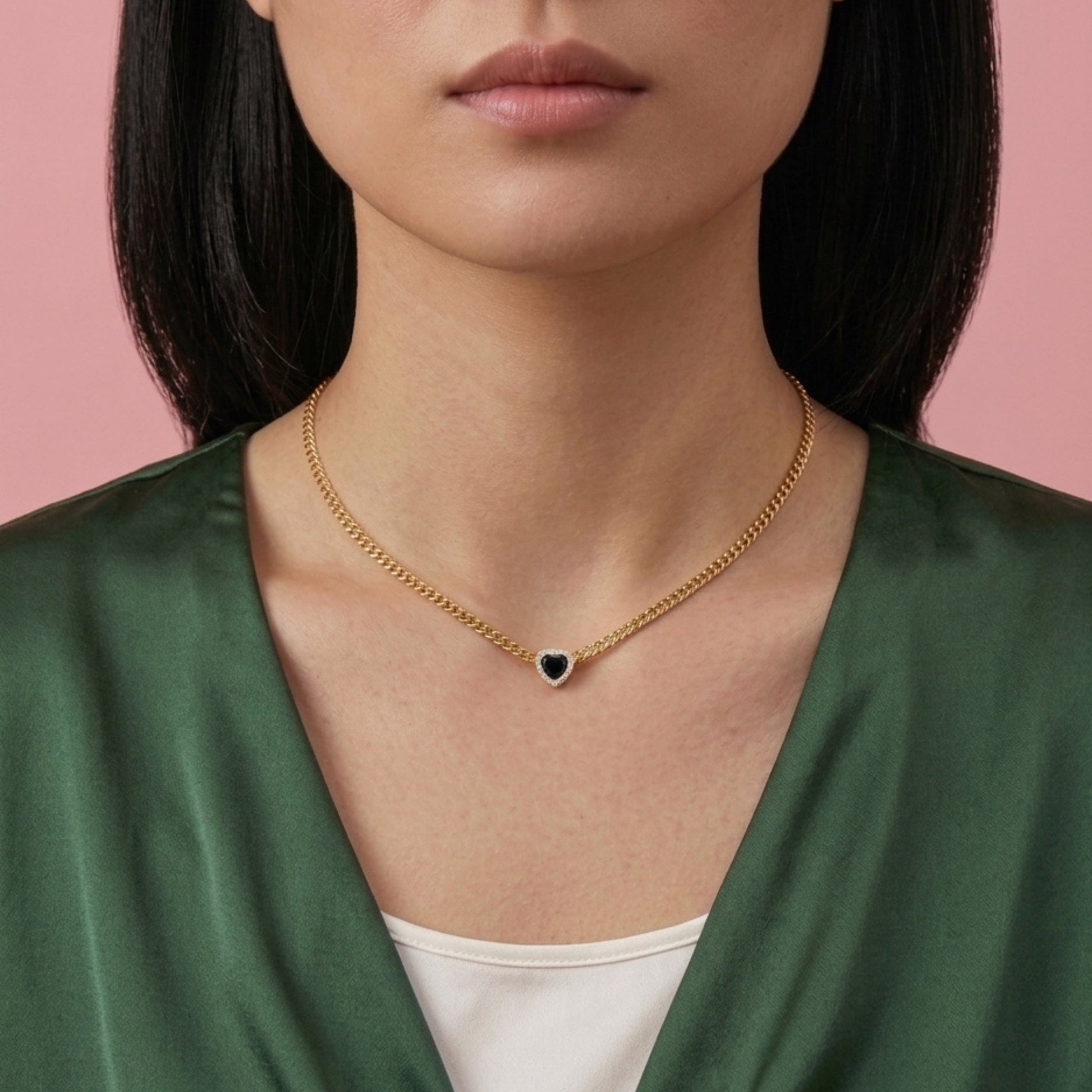 A close-up of a model wearing a delicate gold chain necklace with a small heart-shaped pendant featuring a dark blue center, styled with a green satin top against a solid pink background.