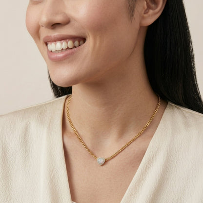 A close-up of a woman wearing a delicate gold chain necklace with a small, round, light-colored stone pendant, styled with a cream-colored top against a soft beige background.