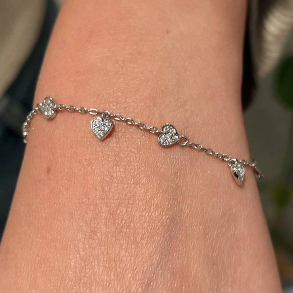 A delicate silver chain bracelet with four small heart charms, each set with clear stones, shown worn on a person's wrist.