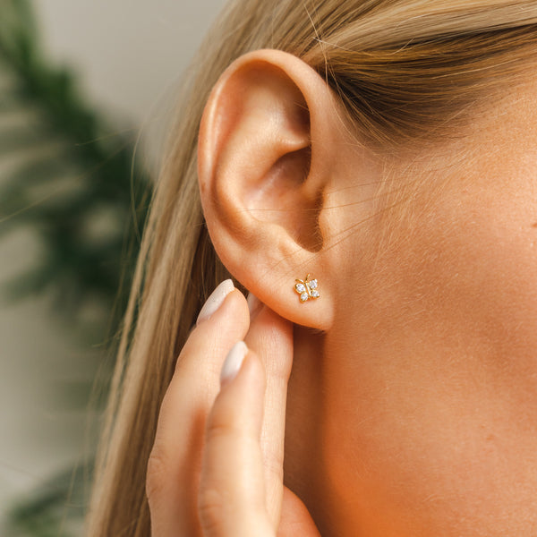 Lifestyle shot of the BLOOMTINE gold butterfly moissanite studs on a woman's ear. These gold-vermiel-over-silver earrings showcase the perfect scale for a dainty yet impactful look, highlighting the tarnish-resistant finish and high-brilliance stones against a soft-focus background
