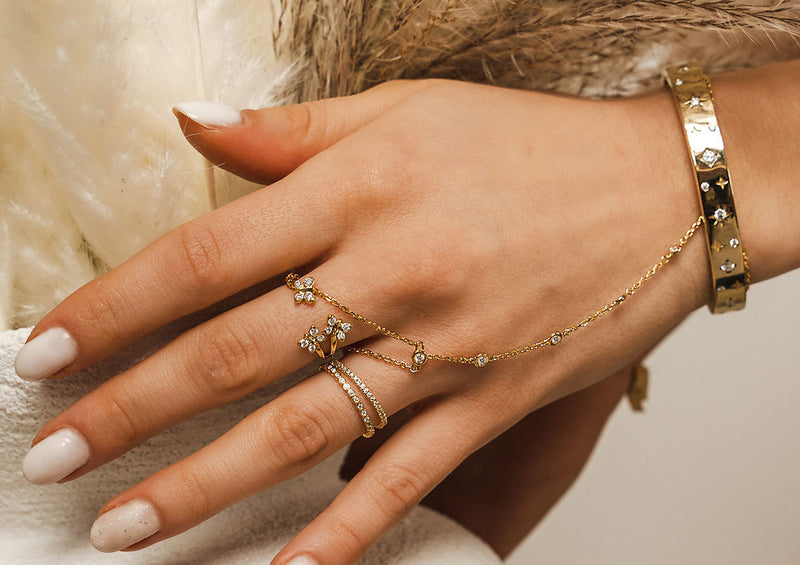Hand wearing gold jewelry with a neutral background. Adjustable sterling silver finger chain by BLOOMTINE with 6 lab-grown diamonds. Known as a hand harness or slave bracelet, this tarnish-resistant rhodium-plated piece features a diamond-cut chain for maximum light refraction. An ethical, high-sparkle luxury gift